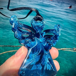 May include: A blue resin angel pendant with glitter accents, suspended from a black cord. The angel has outstretched wings and is held against a backdrop of blue water and sky.