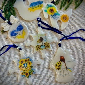 Può includere: Una collezione di ornamenti in resina bianca con nastri blu. Gli ornamenti presentano vari disegni, tra cui una colomba, una capra, un fiocco di neve e una ragazza con girasoli. Alcuni ornamenti hanno testo in ucraino.