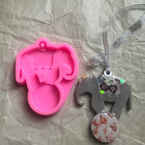 Peut inclure: Un moule en silicone rose en forme d'éléphant avec un anneau, à côté d'un pendentif éléphant gris avec des accents floraux et un globe décoratif. Le pendentif est attaché à un ruban argenté.
