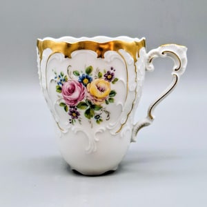 Könnte beinhalten: Weiße Porzellantasse mit goldenem Rand und Henkel. Die Tasse hat ein Blumendesign mit rosa, gelben und blauen Rosen. Der Henkel und die dekorativen Akzente sind ebenfalls goldfarben.