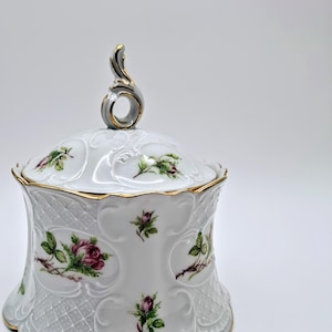 Puede incluir: Tarro con tapa de porcelana blanca con ribete dorado y asa decorativa. El tarro presenta un diseño floral con rosas rosas y hojas verdes. El tarro tiene un patrón en relieve detallado.