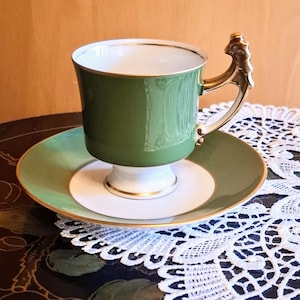 May include: A green and white porcelain teacup and saucer set with a gold trim. The teacup has a unique, ornate handle.