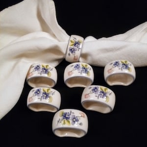 Könnte beinhalten: Set aus sieben Serviettenringen aus Keramik mit Blumenmuster. Die Ringe sind weiß mit blauen und rosa Blumen.