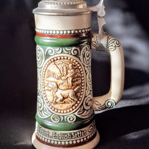 Puede incluir: Una jarra de cerveza de cerámica con un diseño verde y marrón que representa una escena de caza con un perro y una persona. La jarra tiene una tapa de plata con un asa con bisagras y el texto "At Point... English Setter" en la parte inferior.