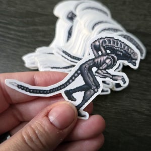 May include: A stack of white stickers featuring a pixelated illustration of an Alien creature in shades of gray and black. The Alien is in a dynamic pose, with its tail extended and arms reaching forward. The stickers are on a dark surface.