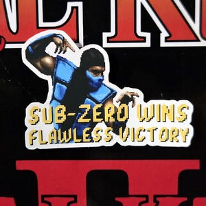 May include: A black background with the words "Mortal Kombat II" in red and orange. A sticker of Sub-Zero in a blue and black outfit is in the center, with the text "Sub-Zero Wins Flawless Victory". Green dragons are on either side.