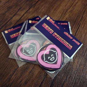 May include: A collection of Death Stranding sticker packs. Each pack contains heart-shaped stickers with a smiling face design, outlined in pink neon. The packs are sealed in clear plastic, with a dark blue header card that reads "DEATH STRANDING" in orange.