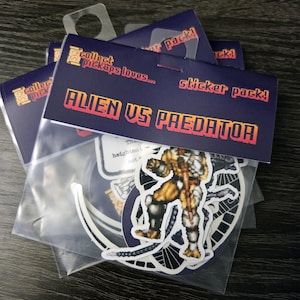May include: A clear plastic bag containing a sticker pack with a header card that reads "collect pickups loves... sticker pack! ALIEN VS PREDATOR". The pack includes pixel art stickers featuring a yellow and brown creature and a dark alien figure.
