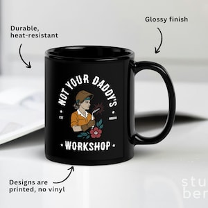 May include: Black ceramic mug with a glossy finish. The mug features the text "NOT YOUR DADDY'S WORKSHOP" and an illustration of a female welder. The design is printed, and the mug is durable and heat-resistant.