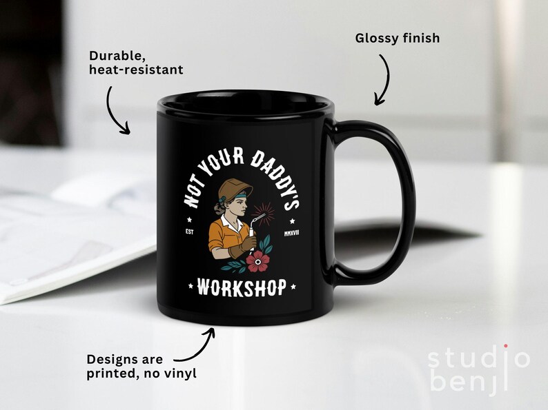 May include: Black ceramic mug with a glossy finish. The mug features the text "NOT YOUR DADDY'S WORKSHOP" and an illustration of a female welder. The design is printed, and the mug is durable and heat-resistant.