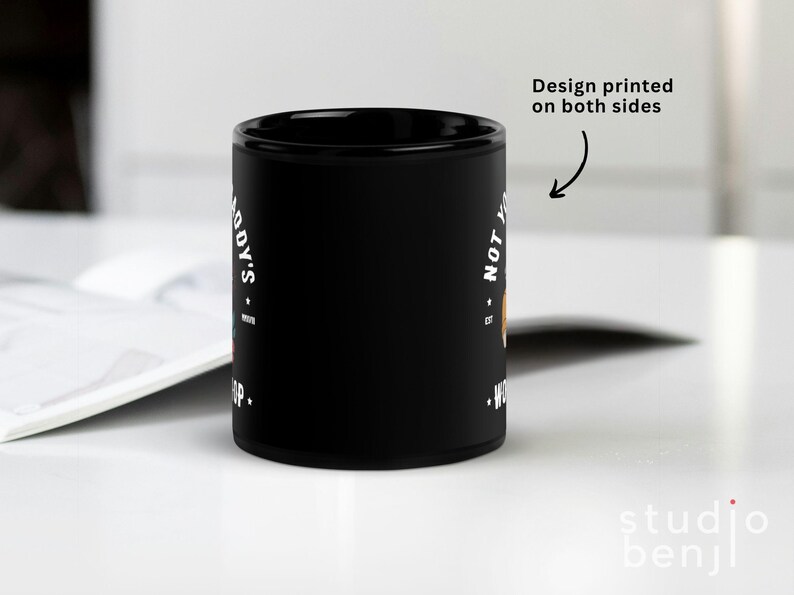 May include: Black ceramic mug with white text that reads "NOT YOUR BODYS WORKSHOP" and "S.A.D.Y'S". The design is printed on both sides. The mug is on a white surface.
