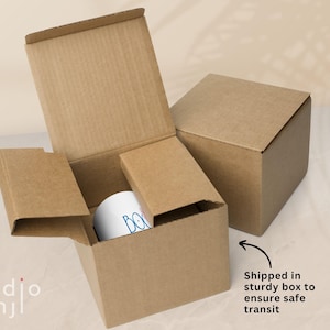 May include: A white mug with blue text inside a cardboard box, with another box in the background. The boxes are brown and the mug is visible inside the open box. The text on the mug is partially visible. The image includes the text "Shipped in sturdy box to ensure safe transit."