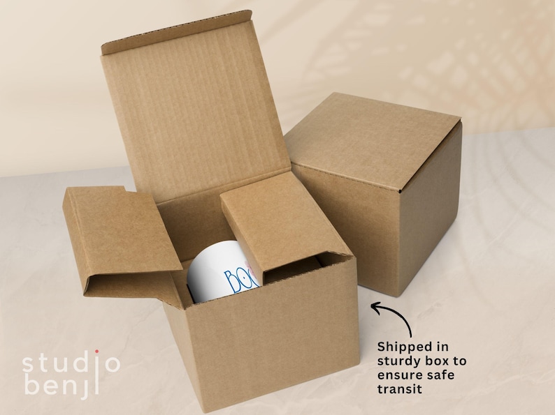May include: A white mug with blue text inside a cardboard box, with another box in the background. The boxes are brown and the mug is visible inside the open box. The text on the mug is partially visible. The image includes the text "Shipped in sturdy box to ensure safe transit."