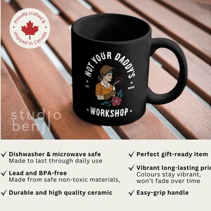 May include: Black ceramic mug with the text "NOT YOUR DADDY'S WORKSHOP" and an illustration of a female welder. The mug has a large handle and is on a wooden surface. The mug is dishwasher and microwave safe.