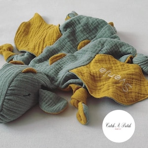 Puede incluir: Un arrullo para bebé con forma de dragón, de color verde azulado y amarillo mostaza. El arrullo tiene una tela arrugada y texturizada y presenta detalles bordados, incluido el nombre "Svenja". El dragón tiene una cara sonriente y una cola anudada.