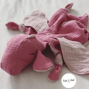 Könnte beinhalten: Ein rosa und hellrosa Stofftier mit einem deckenartigen Körper. Das Plüschtier hat einen Kopf, Ohren und Beine. Das Spielzeug ist aus einem weichen, strukturierten Stoff gefertigt. Das Bild enthält einen weißen Kreis mit dem Text "Catch A Patch" in Schreibschrift.