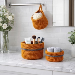 Peut inclure: Ensemble de trois paniers de rangement crochetés orange et gris. L'un est suspendu à une barre métallique, contenant une éponge blanche. Deux autres sont posés sur un comptoir, l'un avec des articles de toilette et l'autre avec des serviettes roulées.