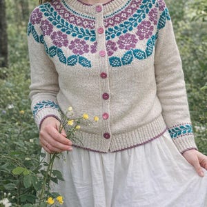 May include: A beige cardigan with a floral pattern in teal and purple. The sweater features a round neck, long sleeves, and pink buttons. The cuffs and neckline are trimmed with a dark purple border. The sweater is worn over a white dress.