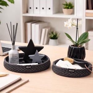 Peut inclure: Deux plateaux ronds en crochet gris foncé sur un bureau en bois. L'un contient un flacon blanc avec des roseaux noirs et deux décorations en forme d'étoile. L'autre contient une brosse et des articles blancs. Un ordinateur portable et un bloc-notes sont également sur le bureau.