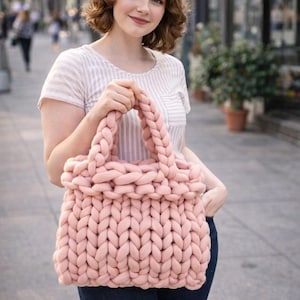 Peut inclure: Un grand sac fourre-tout rose tricoté, doux, avec de larges anses tressées. Le sac est fait de grosse laine et a un aspect texturé. Le sac est tenu par une personne portant un haut rayé et un jean bleu.