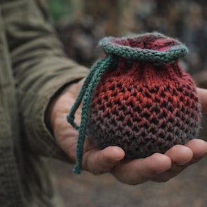 May include: A small, hand-knitted pouch in shades of red, orange, and grey, held in an open hand. The pouch has a green drawstring and trim. The knitted texture is visible, and the background is blurred.