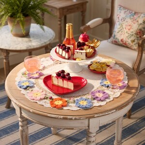 Peut inclure: Une table ronde en bois avec une base blanche, dressée pour un goûter. Elle présente un présentoir à gâteaux blanc avec des tranches de gâteau, une assiette en forme de cœur avec du gâteau et des dessous de verre en forme de fleurs au crochet colorées.