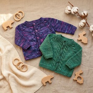 Peut inclure: Deux cardigans pour bébés tricotés, l'un violet avec un motif moucheté et l'autre vert avec un motif torsadé. Des jouets en bois et une couverture crème sont disposés autour des vêtements. Le tout sur un fond neutre.