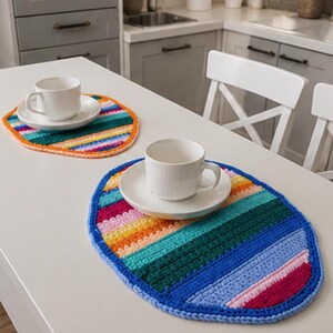 Peut inclure: Deux sets de table ovales en crochet multicolore avec des rayures horizontales dans des tons de bleu, vert, jaune, orange et rose. Chaque set de table supporte une tasse à café en céramique blanche et sa soucoupe assortie.