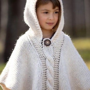 Peut inclure: Un poncho à capuche en tricot crème avec une grande fermeture à bouton en bois. Le poncho présente un motif en tricot torsadé sur le devant et est porté par une jeune personne.