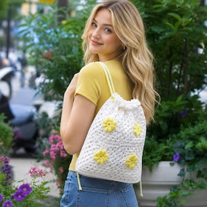 Peut inclure: Un sac à cordon blanc au crochet orné d'appliqués de marguerites jaunes. Le sac a une fermeture à cordon et des bretelles jaunes. Le sac est porté sur l'épaule.