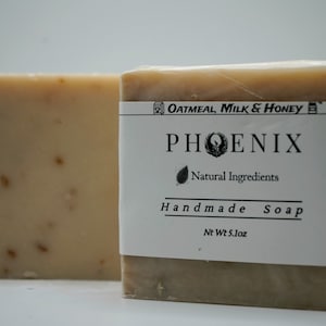 May include: Two rectangular bars of handmade soap. One bar is unwrapped, showing a light brown color with visible flecks. The other bar is wrapped with a label that reads "Oatmeal, Milk & Honey" and "Phoenix Handmade Soap" with a net weight of 5.1 oz.