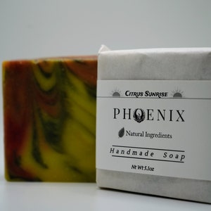 May include: Two bars of handmade soap. One bar is a colourful swirl of yellow, orange, and dark green. The other bar is wrapped in white paper with a label that reads "Citrus Sunrise PHOENIX Natural Ingredients Handmade Soap" and "Net Wt 144.5g."