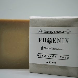 Puede incluir: Dos barras de jabón artesanal. Una barra está sin envolver, mostrando un color marrón claro. La otra barra está envuelta en papel gris con una etiqueta que dice "Cowboy Cologne PHOENIX Natural Ingredients Handmade Soap" y Peso Neto 144g.
