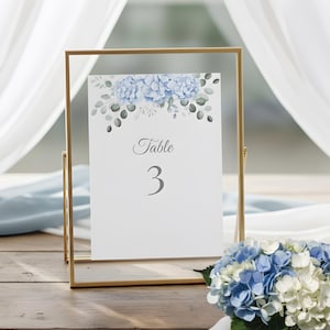 May include: A gold metal framed table number card with a floral design. The card displays the word "Table" and the number "3" in gray script. A bouquet of blue and white flowers sits in front of the card.