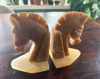 May include: A pair of horse head bookends carved from a light brown stone. The bookends are in the shape of a horse's head and neck, with a rectangular base. The horse's mane is detailed with vertical lines.