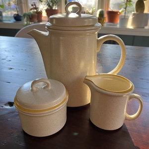May include: A cream-colored ceramic coffee pot with a yellow trim, a matching creamer, and a sugar bowl with a lid. The set is on a wooden table.