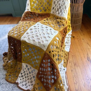 May include: A handmade crochet blanket draped over a chair. The blanket features a patchwork design with squares in shades of mustard yellow, brown, and cream. The openwork pattern adds texture and visual interest to the cozy home decor item.