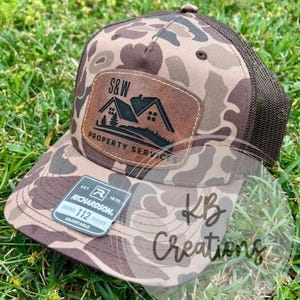 May include: A brown and tan camouflage trucker hat with a brown leather patch that says "S&W Property Services". The hat is adjustable and has a black mesh back.