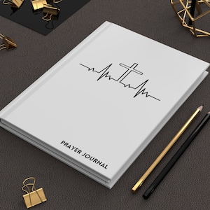 Op de afbeelding: Wit gebedsdagboek met een zwart kruis en een hartslagontwerp. Het dagboek heeft de tekst "PRAYER JOURNAL" op de voorkant.