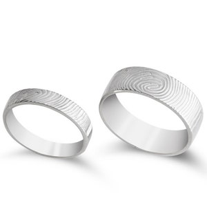 May include: Two silver-toned rings with a polished finish, each featuring a distinct, engraved fingerprint pattern wrapping around the entire outer band. The rings are displayed side-by-side, showcasing the detailed, swirling texture of the metal.