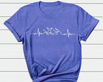 Könnte beinhalten: Ein blaues T-Shirt mit einem weißen Grafikdruck von einer Bergkette und Vögeln. Die Grafik ist ein stilisierter Herzschlag mit Bergen und Vögeln, die darüber fliegen.