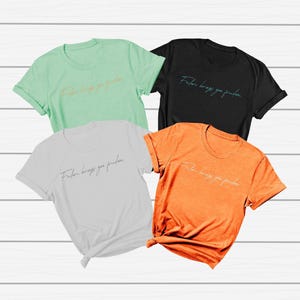 Pode incluir: Quatro camisetas de manga curta em cores diferentes: verde menta, preto, branco e laranja. Cada camisa tem o texto "Faith brings you freedom" em fonte cursiva. As camisas são exibidas em uma superfície de madeira branca.