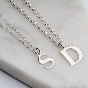 May include: Two silver necklaces featuring polished letter pendants. One necklace has a capital letter S pendant, and the other necklace has a larger capital letter D pendant. Both pendants are attached to matching silver rolo chains with round links.