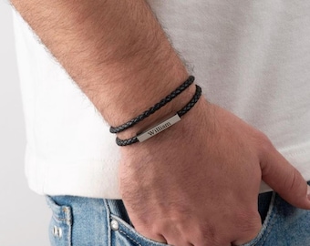 May include: Black braided leather bracelet with a silver bar engraved with the name "William". The bracelet is worn on a wrist, with a second strand of the bracelet visible. The person is wearing a white shirt and blue jeans.