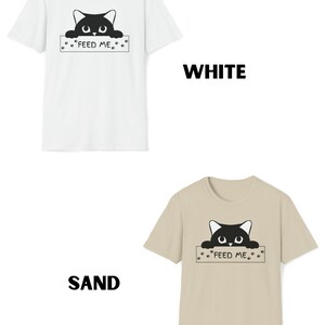May include: Two t-shirts with a black cat peeking over a sign that says "Feed Me". The t-shirts are available in white and sand.
