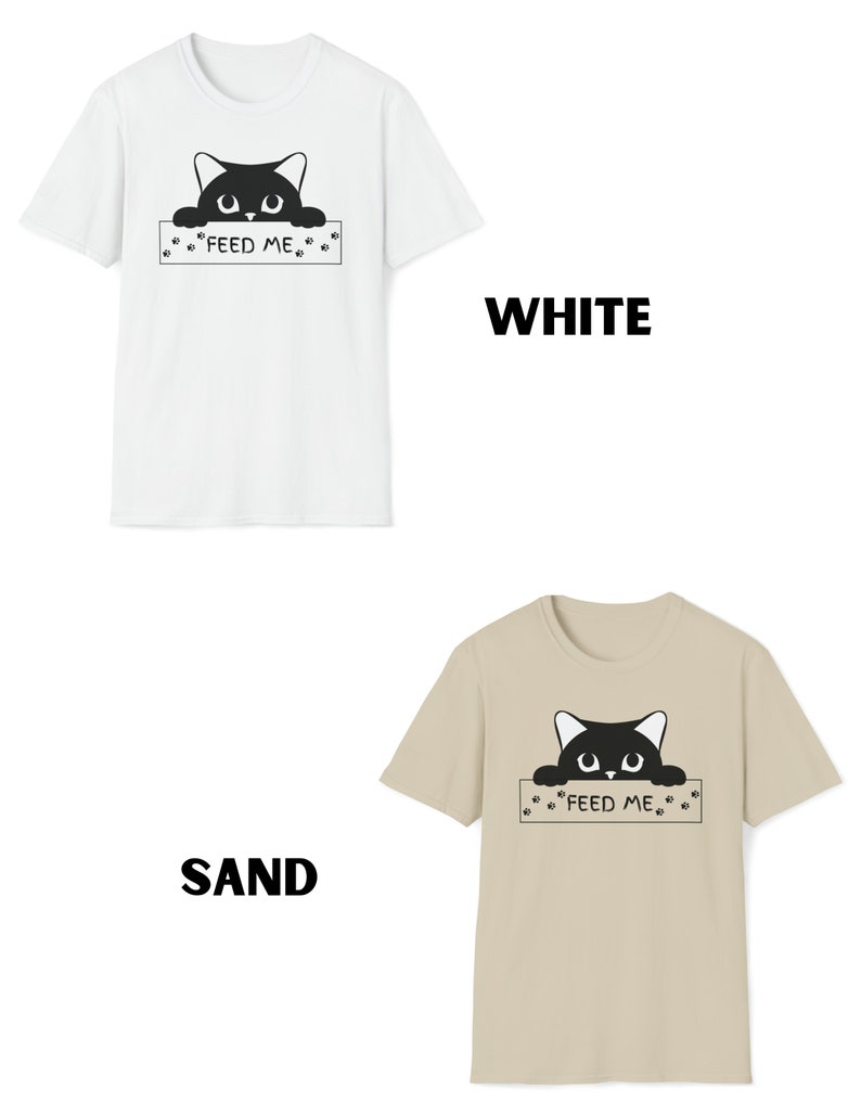 May include: Two t-shirts with a black cat peeking over a sign that says "Feed Me". The t-shirts are available in white and sand.
