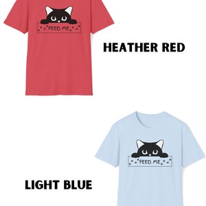 May include: Two t-shirts with a black cat peeking over a sign that says "Feed Me". The t-shirt on the left is heather red and the t-shirt on the right is light blue.