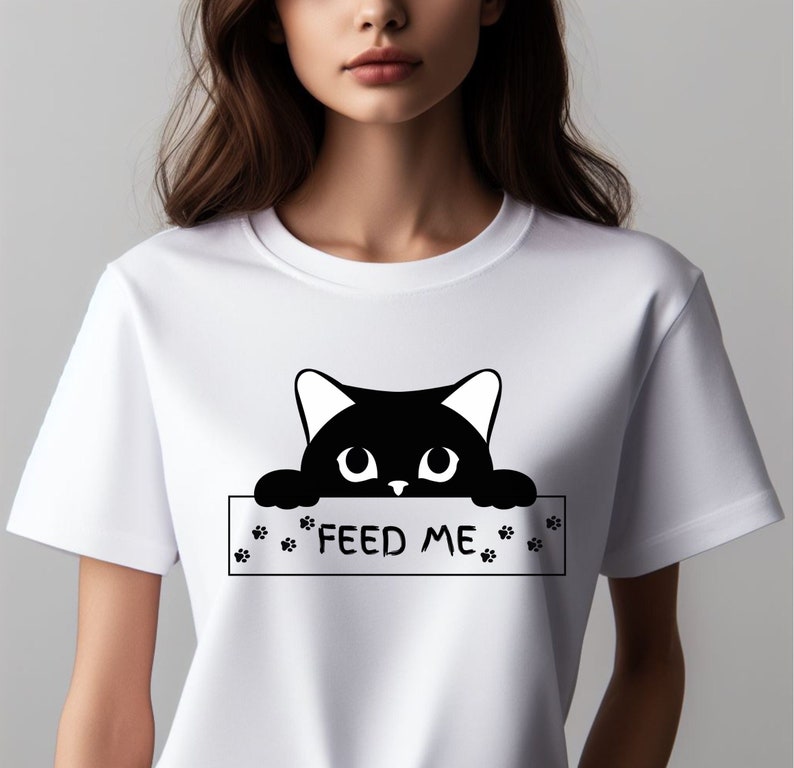 May include: White t-shirt with a black cat peeking over a sign that says "FEED ME". The sign has paw prints on either side.