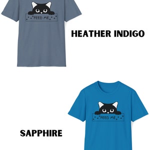 May include: Two t-shirts with a black cat peeking over a sign that says "Feed Me". The t-shirt on the left is heather indigo and the t-shirt on the right is sapphire.