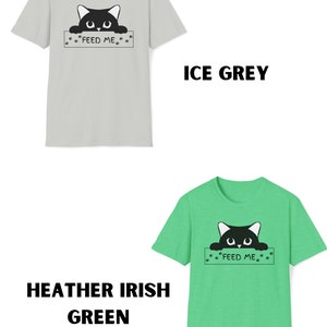 May include: Two t-shirts with a black cat peeking over a sign that says "Feed Me". The shirt on the left is light gray and the shirt on the right is a light green heather.